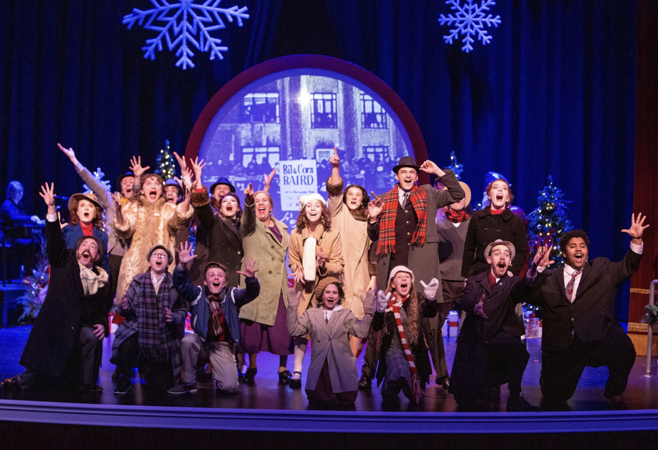 NUOVA Vocal Arts performers onstage in Miracle on 34th Street beneath winter snowflakes and a shop-window backdrop.